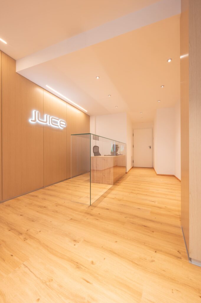 Desk reception in vetro e legno chiaro con insegna luminosa al neon "JUICE" a parete e pavimento in parquet.