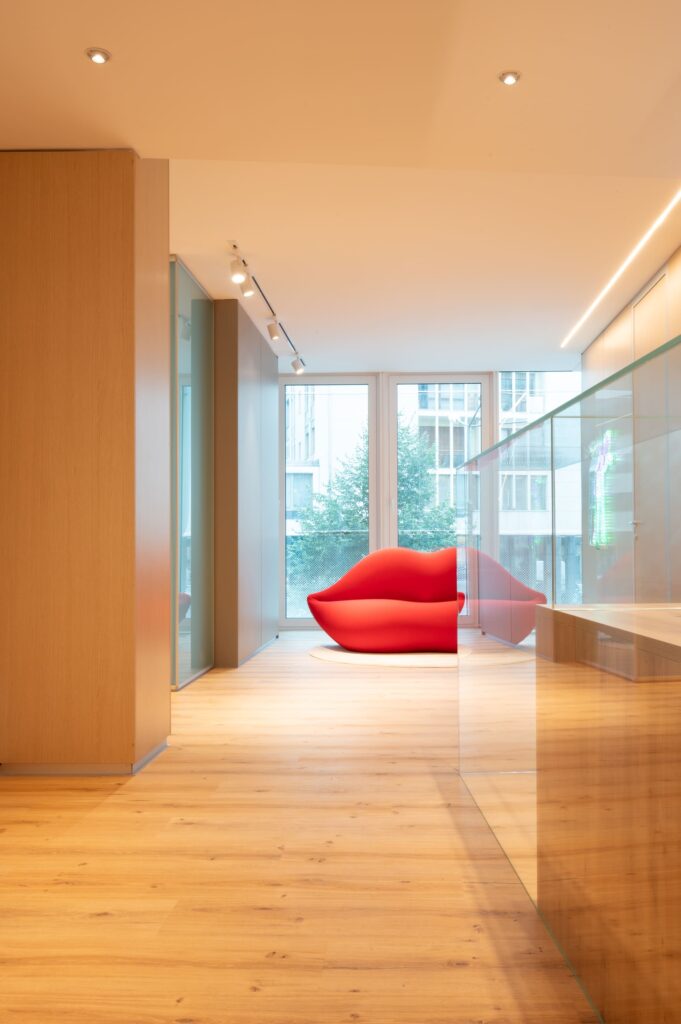 Corridoio d'ufficio moderno con pavimento in parquet e vista su divano rosso a forma di labbra posizionato di fronte a una vetrata.