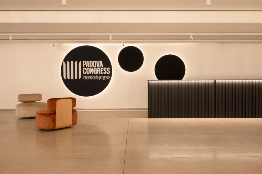 Area accoglienza con bancone scuro a doghe, poltroncine di design e pannelli acustici circolari neri a parete con brand Padova Congress.