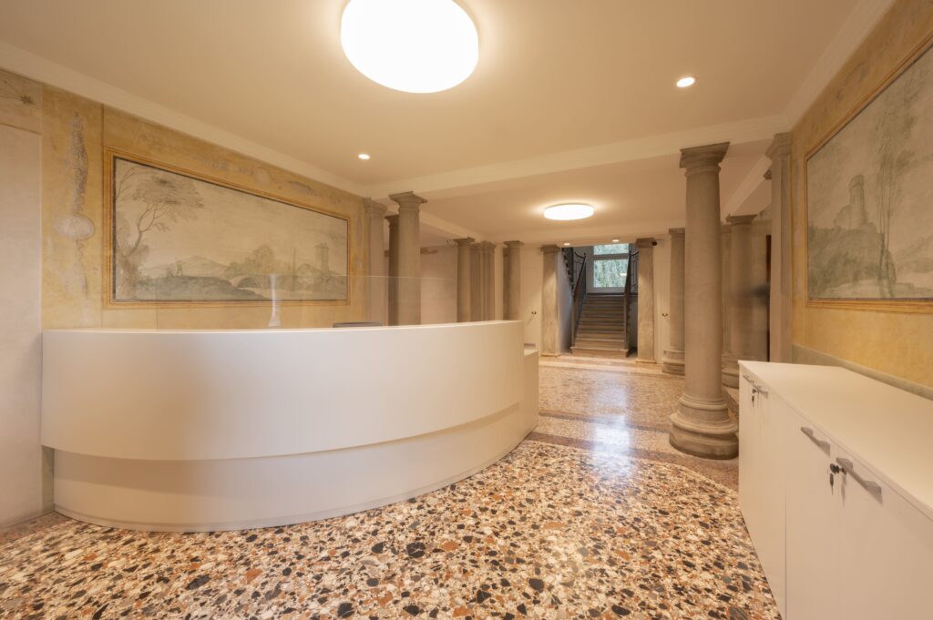 Hall d'ingresso d'epoca con pavimento in terrazzo veneziano, colonne in pietra, affreschi e moderno bancone reception curvo bianco.