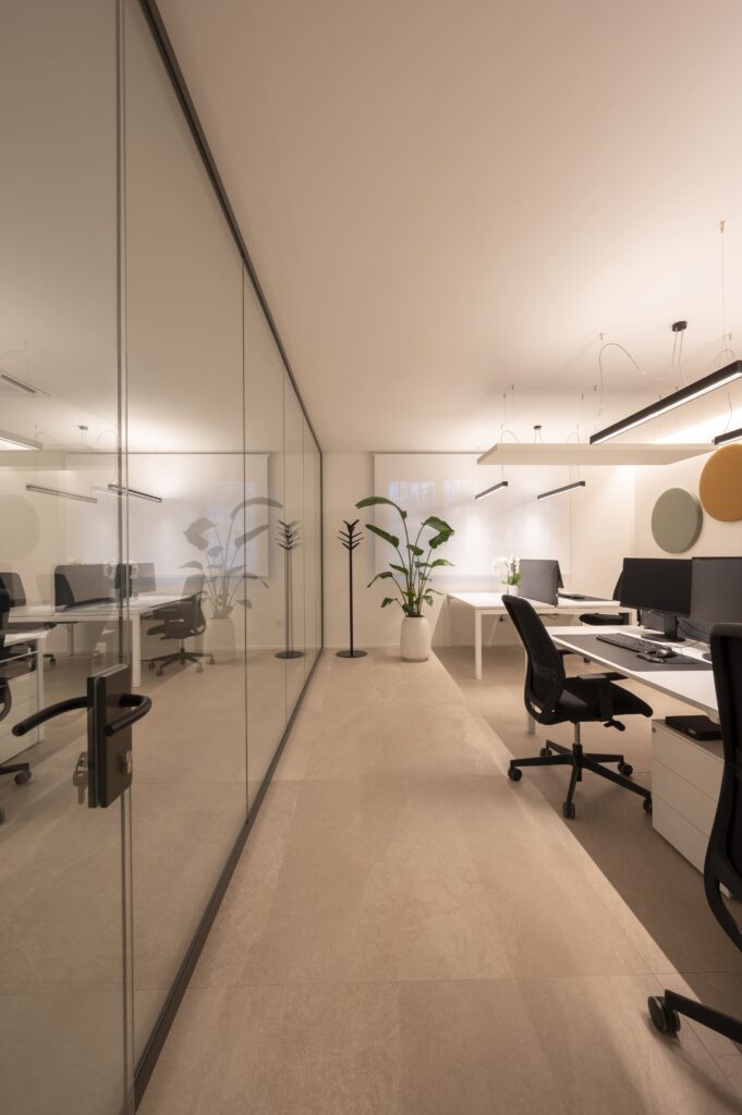 Corridoio di un ufficio moderno delimitato da pareti divisorie in vetro e postazioni operative con luci a LED.