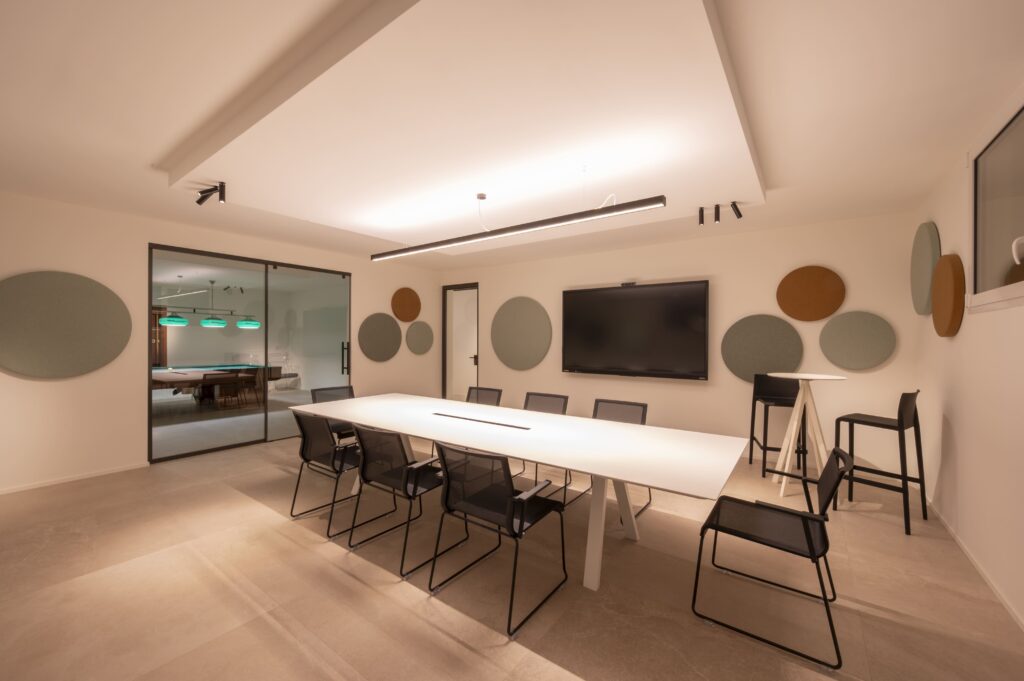 Sala riunioni moderna con lungo tavolo da conferenza bianco, monitor a parete, pannelli acustici circolari e sedie nere.