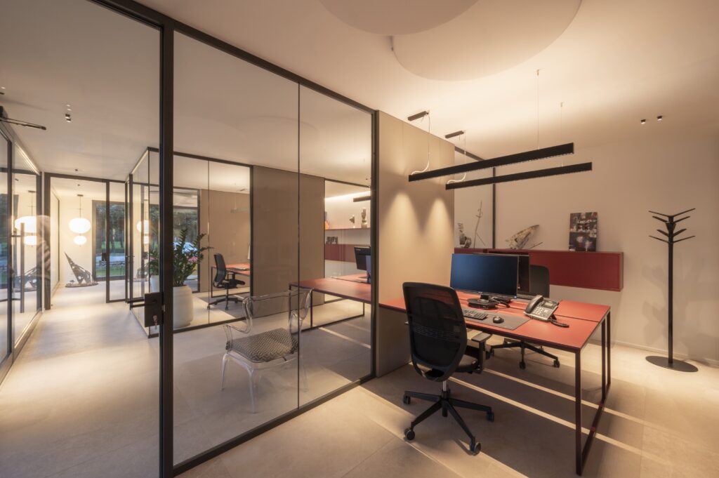 Ufficio delimitato da pareti in vetro con scrivania bordeaux, PC, appendiabiti nero e sedia trasparente di design.