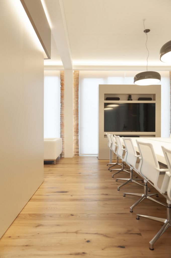 Sala riunioni con lungo tavolo bianco, sedie ergonomiche a rete, parquet e mobile con TV integrata.